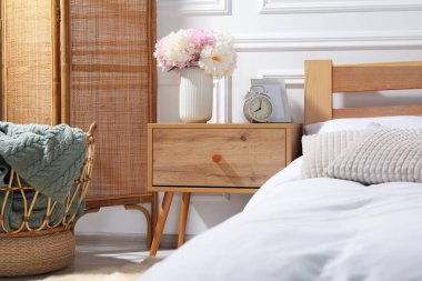 Komodinin yanında çalar saat ve yatağın yanında çiçekler var.