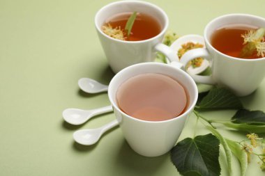 Fincanlarda tatlı ıhlamur çayı, kaşıklar, yapraklar ve açık yeşil arka planda çiçekler, yakın plan. Metin için boşluk