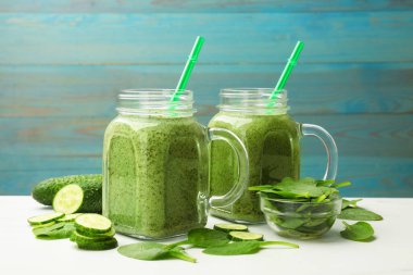 Mason kavanozlarında lezzetli bir smoothie, beyaz masada ıspanak yaprakları ve salatalık açık mavi ahşap arka planda, yakın plan