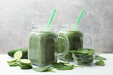 Mason kavanozlarındaki lezzetli smoothie, beyaz masada ıspanak yaprakları ve salatalık, gri arka planda, yakın planda.