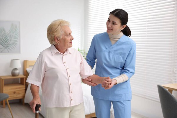 Nurse assisting senior woman with walking cane indoors. Home health care service