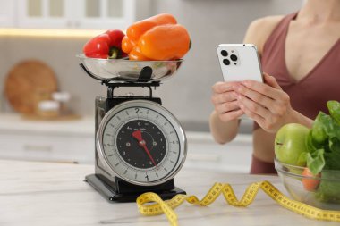 Beyaz mermer masanın yanında akıllı telefon kullanan bir kadın. Mutfak terazisi, sağlıklı yiyecekler ve kapalı alanda mezura kullanıyor.