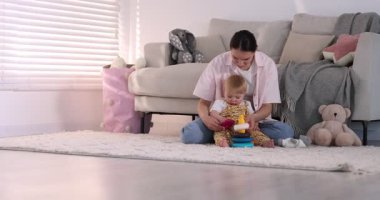 Anne ve küçük bebeği evde oyuncak piramitlerle oynuyorlar, kamera içeri giriyor.