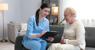 Hemşire elinde dosya ile kanepede yaşlı kadını muayene ediyor. Ev sağlık hizmetleri