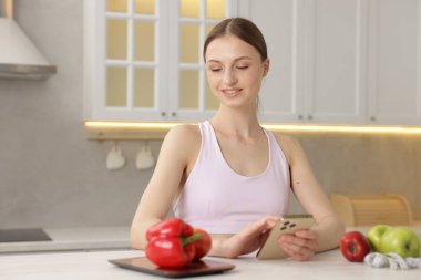 Beyaz masada akıllı telefon kullanan bir kadın. Mutfak terazisi, sağlıklı yiyecekler ve kapalı alanda mezura kullanıyor.