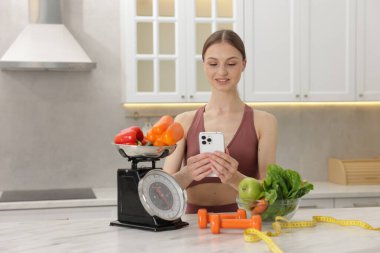 Beyaz mermer masanın yanında akıllı telefon kullanan bir kadın. Mutfak terazisi, sağlıklı yiyecekler, dambıllar ve kapalı alanda mezura kullanıyor.