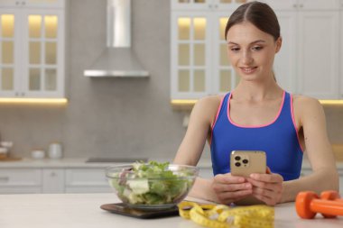 Beyaz masada akıllı telefon kullanan bir kadın mutfak tartısı, salata, mezura ve dambıl kullanıyor.