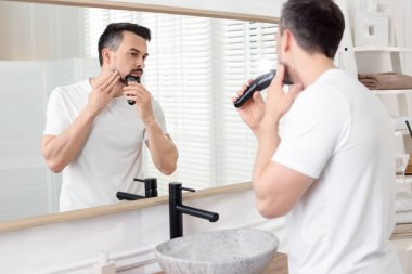 Yakışıklı adam evde aynanın yanında tıraş makinesiyle tıraş oluyor.