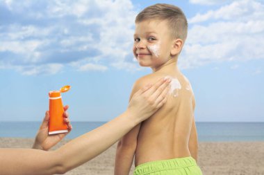 Anne sahilde oğlunun tenine güneş kremi sürüyor, yakın plan. Yazın dışarıda dinlenirken güneş koruması