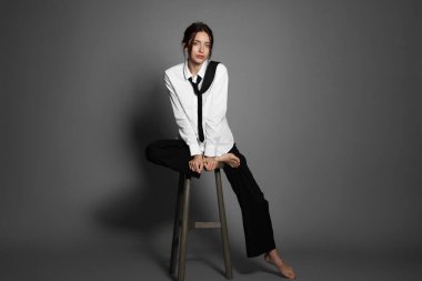 Şık giyinmiş güzel bir kadın açık gri arka planda sandalyede oturuyor.