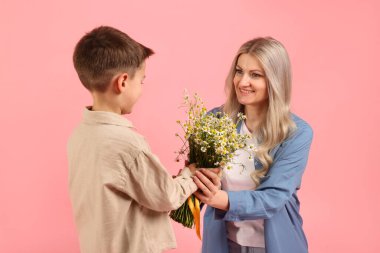 Küçük çocuk, gülümseyen annesine pembe arka planda bir buket güzel papatya çiçeği sunuyor.