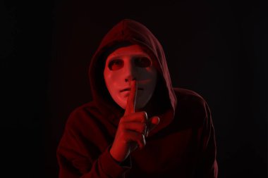 Anonimlik. Maskeli adam karanlık arka planda kırmızı ışıkta sessiz jest yapıyor.