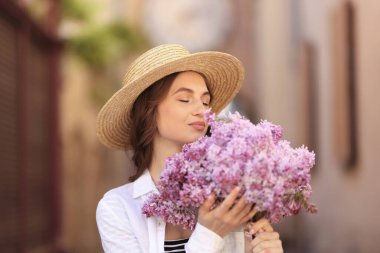 Bulanık arka planda bir demet leylak çiçeğiyle şapkalı güzel bir kadın.