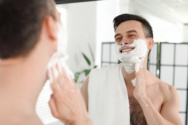 Gülümseyen adam banyoda aynanın yanında tıraş köpüğü sürüyor.