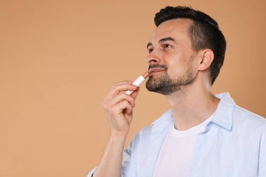 Açık kahverengi arka planda dudaklarına dudak kremi süren yakışıklı bir adam. Metin için boşluk