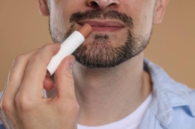 Açık kahverengi arka planda dudaklarına dudak kremi süren adam, yakın plan.