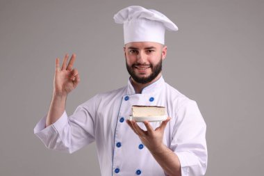 Üniformalı mutlu pastacı elinde lezzetli bir cheesecake tutuyor ve açık gri arka planda güzel bir jest yapıyor.