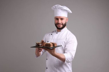 Üniformalı mutlu pastacı açık gri arka planda lezzetli çikolatalı tatlılar tutuyor.