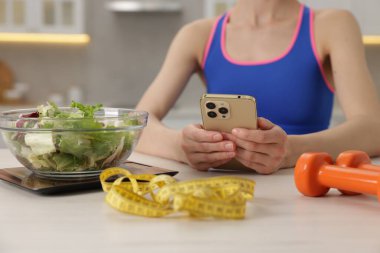 Beyaz masada akıllı telefon kullanan kadın mutfak terazisi, salata, mezura ve halterler kapalı alanda, yakın plan.
