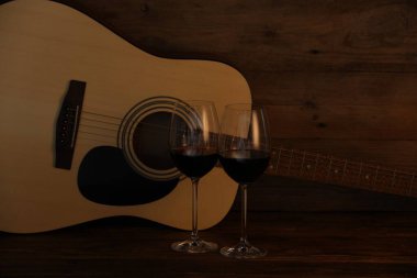 Akustik gitar ve kırmızı şarap kadehleri ahşap masada.