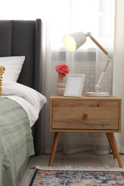 Yatağın yanında lamba, çiçek ve çerçeve olan komodin.