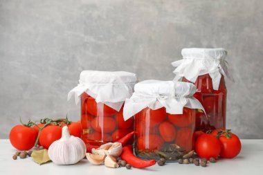 Kavanozlarda domates turşusu, taze ürünler ve baharatlar beyaz masada gri arka planda, yakın planda.