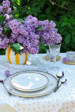 Bahçedeki masada güzel leylak çiçekleri, tabaklar, çatal bıçaklar, bardaklar ve mumlar