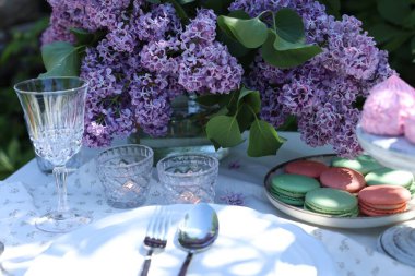 Güzel leylak çiçekleri, tatlılar, tabaklar, çatal bıçak takımı, cam ve yanan mumlar bahçede, yakın plan