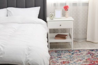 Yatağın yanında çalar saati, çiçeği ve çerçevesi olan bir komodin.