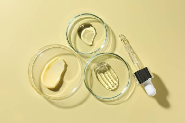 Petri kaplarında farklı kozmetik ürünleri ve soluk sarı arka planda damlalar.