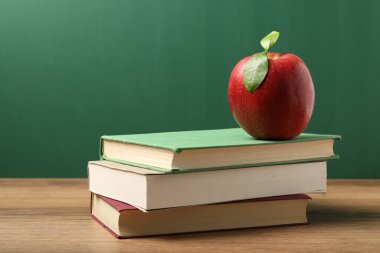 Tahtanın yanındaki ahşap masada farklı kitaplar ve kırmızı elma, yakın plan.