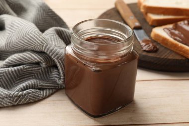 Cam kavanozda çikolatalı tereyağı, açık tahta masada ekmek ve bıçak dilimleri, yakın plan.