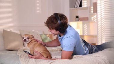 Kulaklıklı, akıllı telefonlu, evde yatakta köpeğini okşayan gülümseyen genç bir çocuk.