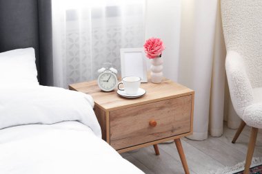 Yatağın yanında kahve, çiçek ve çalar saat olan komodinin yanında.