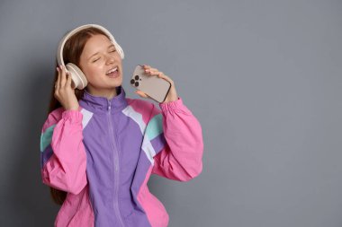 Kulaklıklı, akıllı telefonlu, gri arka planda şarkı söyleyen mutlu genç kız. Metin için boşluk