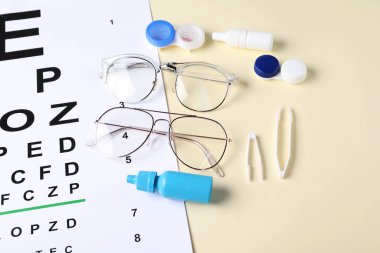 Görüş testi tablosu, gözlükler, cımbızlar, lensler ve bej arka planda tıbbi damlalar.