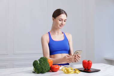 Beyaz mermer masada akıllı telefon kullanan bir kadın mutfak tartısı, sebzeler ve kapalı alanda mezura kullanıyor.