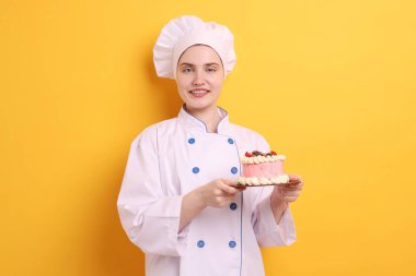 Üniformalı pastacı turuncu arka planda lezzetli bir pasta tutuyor.