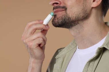 Açık kahverengi arka planda dudaklarına dudak kremi süren adam, yakın plan.