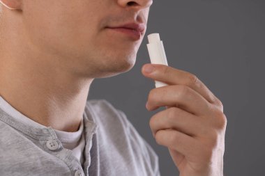 Gri arka planda dudaklarına dudak kremi süren genç adam, yakın plan.