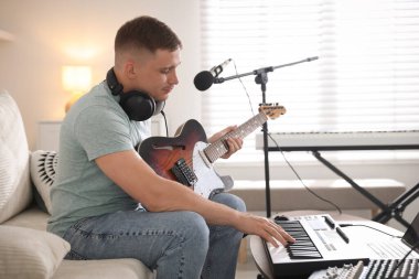 Ev stüdyosundaki kanepede gitar ve klavyeyle çalışan bir adam.