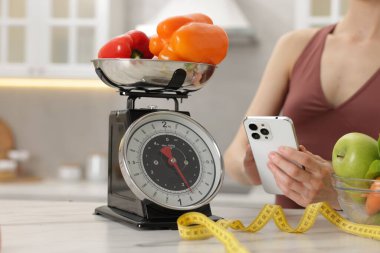 Beyaz mermer masanın yanında akıllı telefon kullanan bir kadın. Mutfak terazisi, sağlıklı yiyecekler ve kapalı alanda mezura kullanıyor.