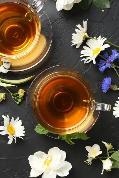 Cam fincanlarda aromatik bitki çayı, farklı çiçekler ve siyah masada nane şekeri.