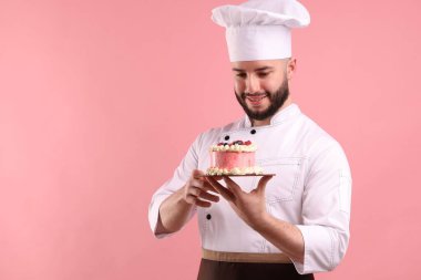 Üniformalı mutlu pastacı pembe arka planda böğürtlenli pasta tutuyor.