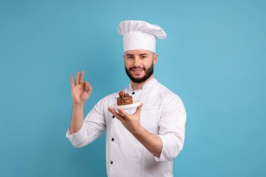 Üniformalı mutlu pastacı elinde çikolatalı tatlı ve açık mavi arka planda iyi bir jest gösteriyor.