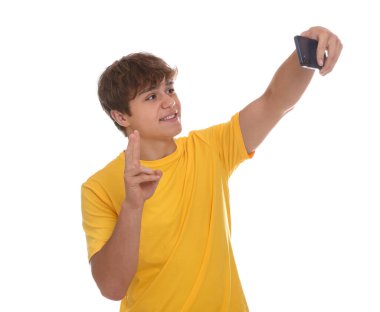 Gülümseyen genç çocuk akıllı telefonuyla selfie çekiyor ve beyaz arka planda barış işareti gösteriyor.