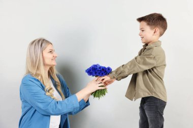 Küçük çocuk açık gri arka planda gülümseyen annesine bir buket güzel çiçek sunuyor.