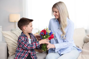Evdeki kanepede gülümseyen annesine bir buket güzel çiçek sunan küçük çocuk.