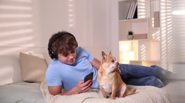 Genç çocuk evde akıllı telefon kullanıyor ve yatakta köpeğini okşuyor.