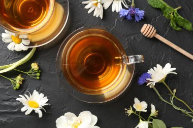 Cam fincanlarda aromatik bitki çayı, farklı çiçekler ve siyah masada nane şekeri.